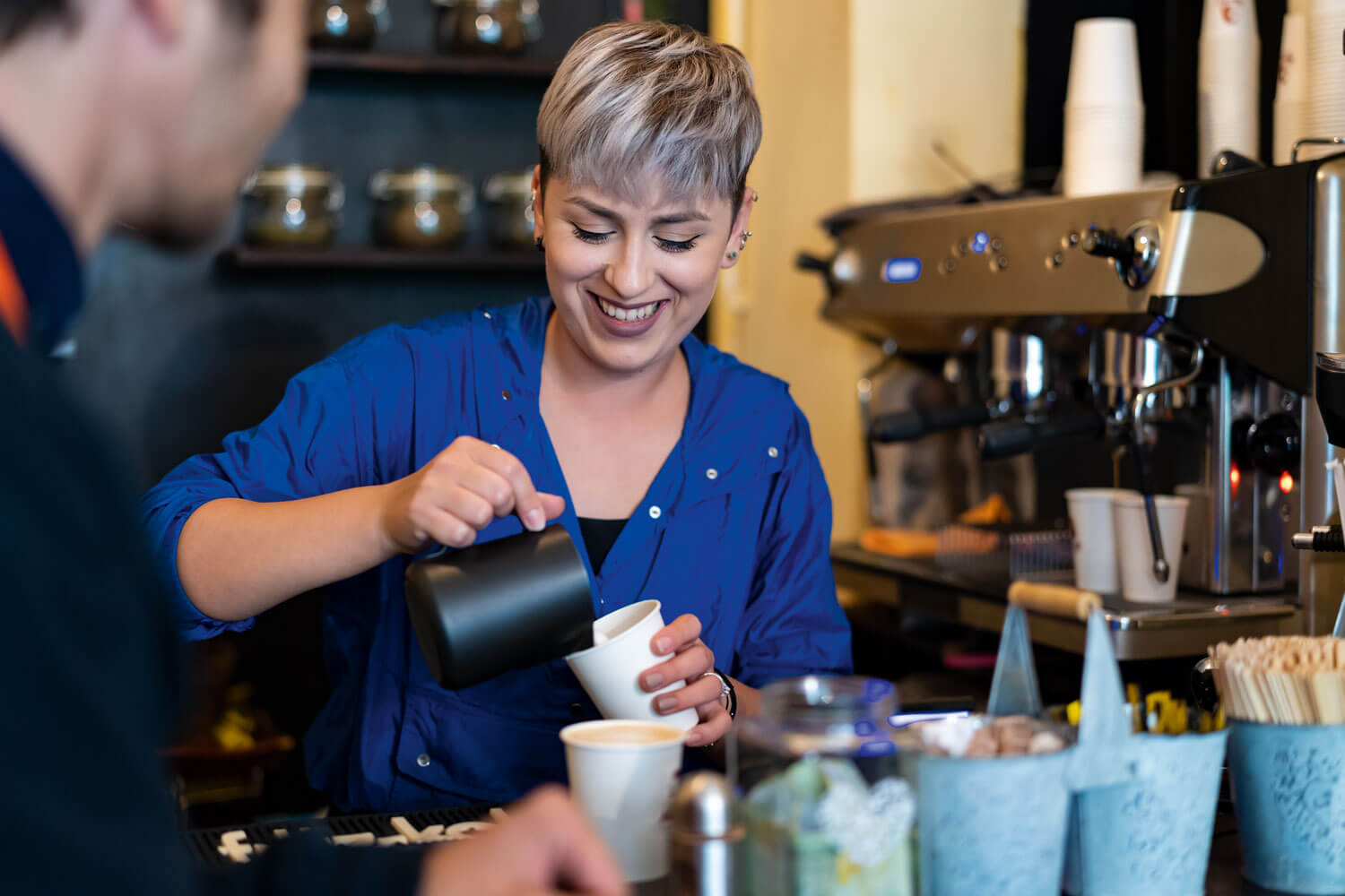 o femeie blondă, tunsă scurt, pregătește o cafea cu lapte într-o cafenea